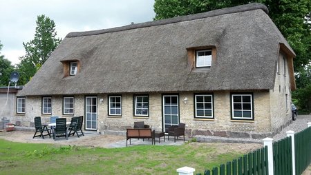 Reetdachhaus mit zwei kleinen Gauben. Heller Stein mit grün-weißen Fenstern und Türen. Davor eine Terrasse mit Gartenmöbeln. 